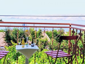 A terrace with a table set for dining, surrounded by metal railings and chairs, overlooking a body of water and greenery.