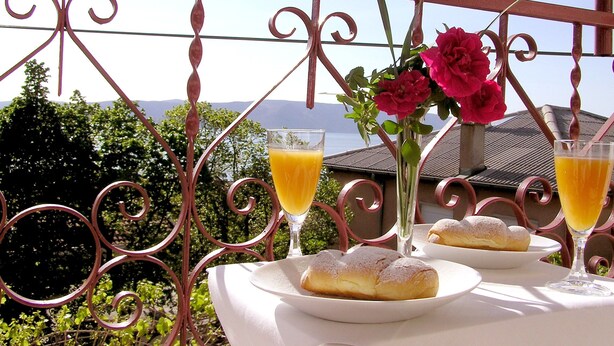 Un balcón con una mesa dispuesta para el desayuno, que incluye bollería y vasos de zumo de naranja, con vistas a un paisaje pintoresco con montañas y una masa de agua.