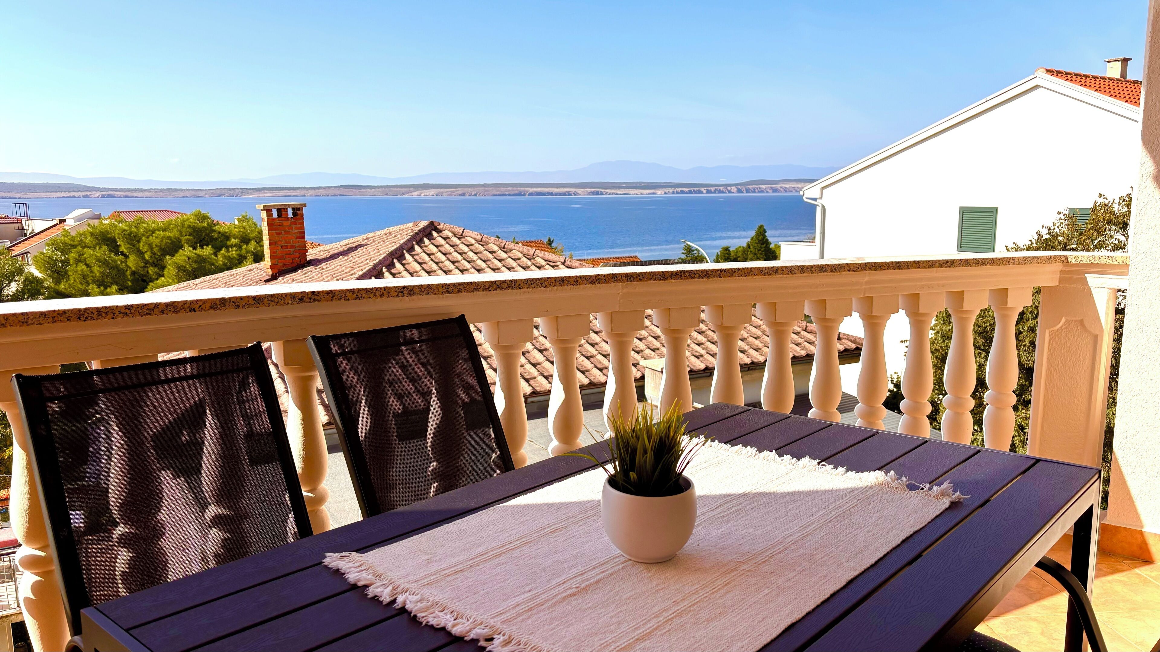 A balcony with a table and chairs, a potted plant, and a view of the sea and distant mountains.