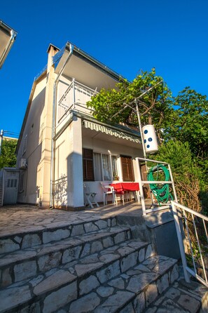 A two-story house with a balcony, stone steps leading to the entrance, and a red table and chair outside.