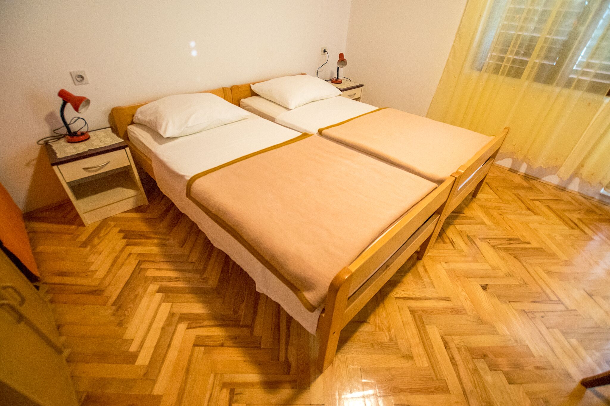 Un lit double avec un cadre en bois et une couvre-lit beige, deux tables de chevet avec des lampes et un parquet en bois à motifs chevrons.