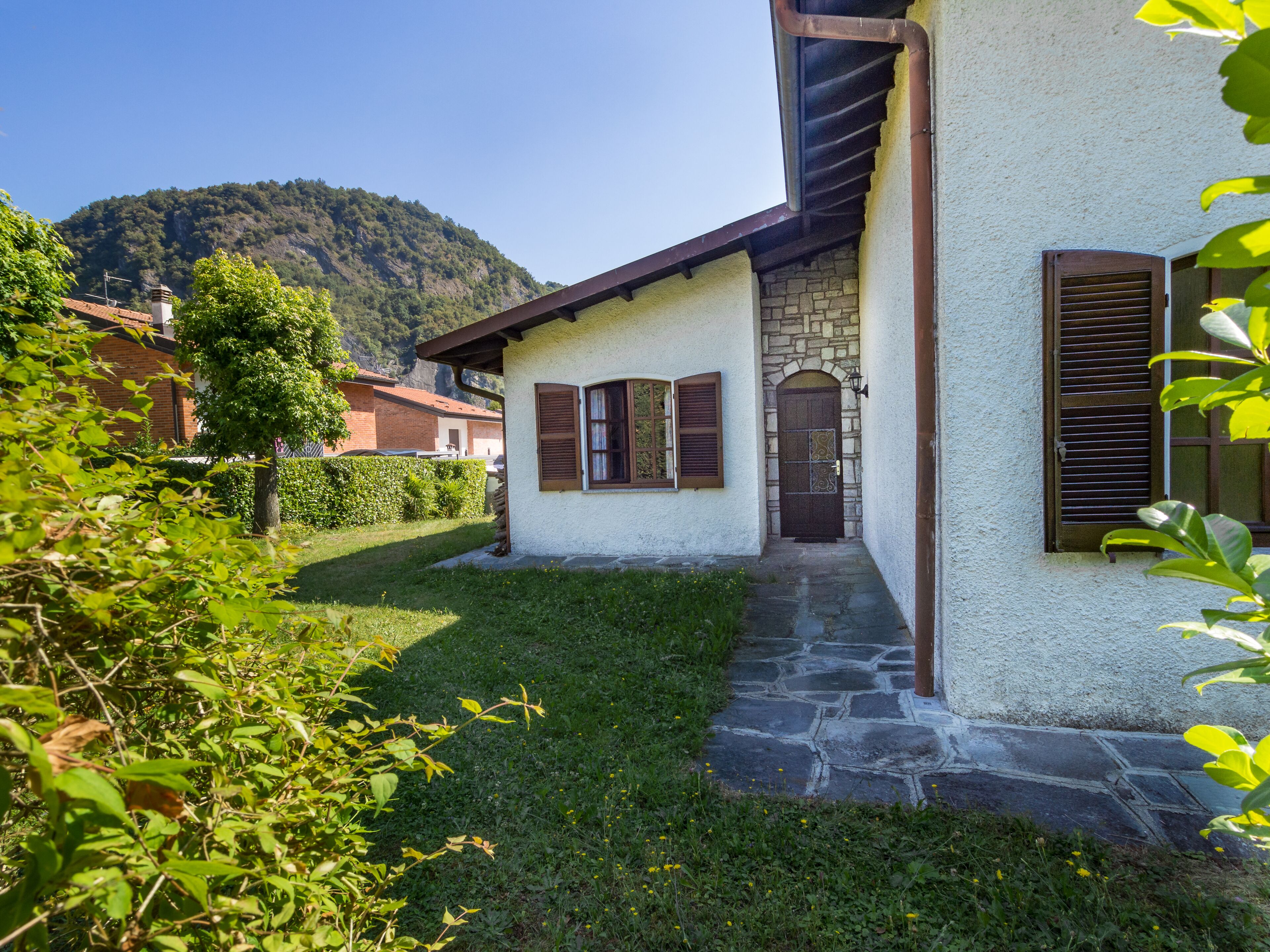 A house with a stone staircase leading to a wooden door, surrounded by greenery and a mountain in the background.