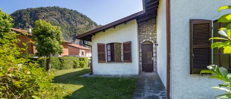 A house with a stone staircase leading to a wooden door, surrounded by greenery and a mountain in the background.