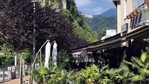 A hotel with a mountain backdrop, a balcony with orange tiles, and a fenced outdoor area with plants. - Spacious vacation home with garden, approx. 150 m to the lake, with bathing facilities (Lavena Ponte Tresa)