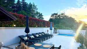 A rooftop pool area with lounge chairs and umbrellas, surrounded by greenery and a clear sky.