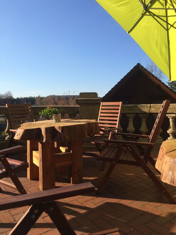 Eine Terrasse im Freien mit Holzmöbeln, darunter ein Tisch und Stühle, und einem gelben Sonnenschirm, der Schatten spendet.