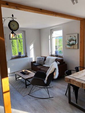 Ein Wohnzimmer mit braunem Sofa, einem schwarzen Schaukelstuhl, einem Holztisch und einem Esstisch. An der Wand hängt eine Uhr und über dem Sofa ein Gemälde.