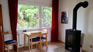 A cozy room with a wooden table and chairs, a window with red curtains, a hanging lamp, and a black wood stove. - Holiday house in the Lüneburg Heath up to 4 people (Suderburg)