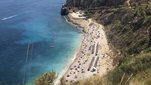 Una spiaggia costiera con una scogliera rocciosa, mare cristallino e numerosi ombrelloni.