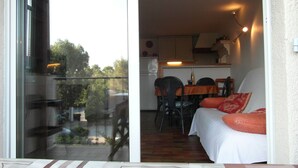 Ein Balkon mit Glastür, eine Essecke mit Tisch und Stühlen, ein Bett mit Kissen sowie eine Küchenzeile mit Schränken und Kühlschrank.