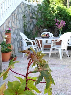 Uma área de pátio com parede de pedra, cadeiras brancas, uma mesa com uma tigela de frutas e plantas em vasos.