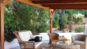 Un espacio cubierto con patio amueblado con muebles de mimbre, incluyendo un sofá y dos sillones, y una mesa redonda de mimbre con un cuenco de fruta.