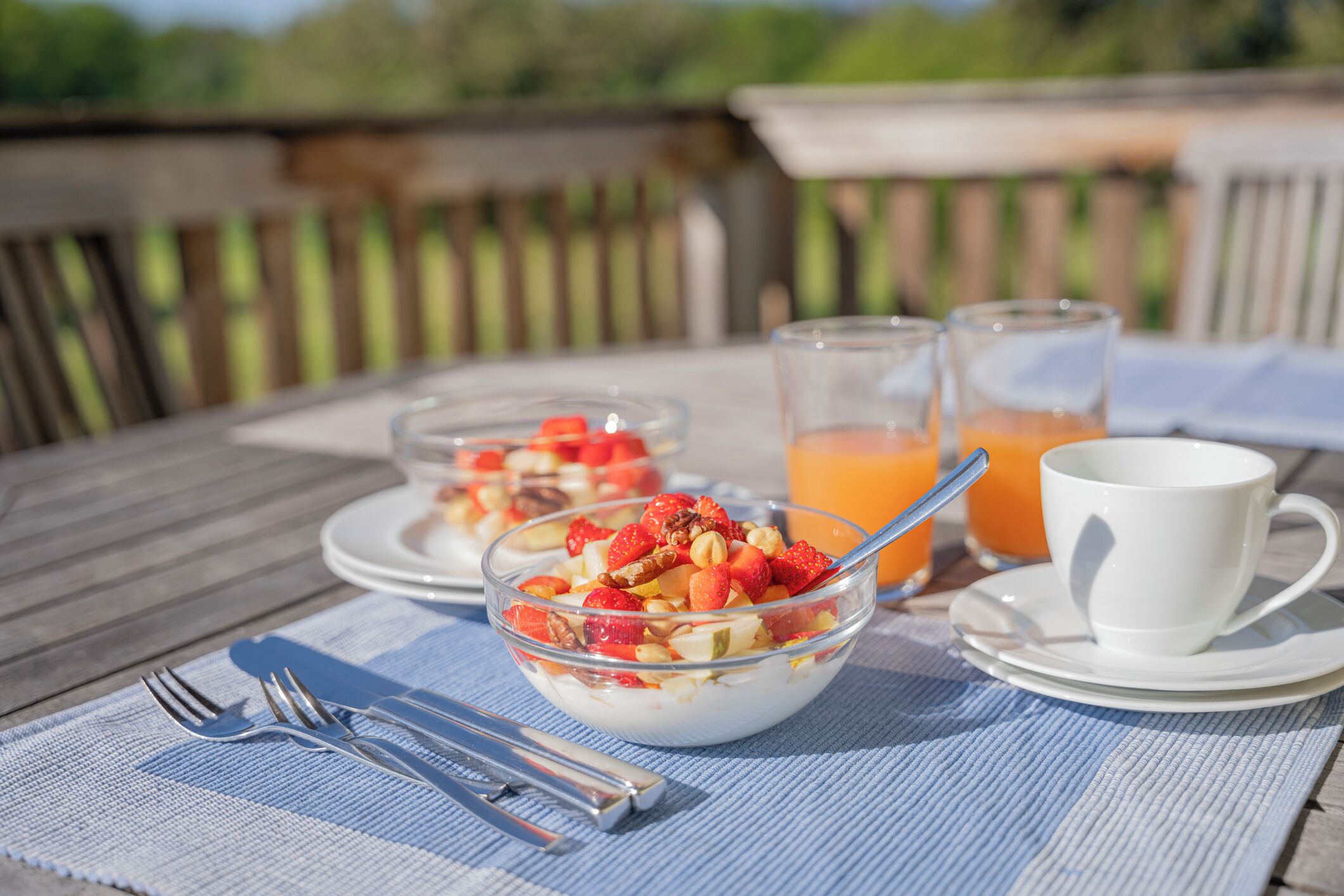 Une table dressée pour le petit-déjeuner, avec un bol de yaourt garni de fruits et de noix, un verre de jus d’orange et une tasse blanche posée sur une table en bois, à l’extérieur.