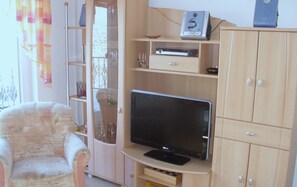 Un salon avec un téléviseur posé sur un meuble en bois, un fauteuil et un buffet avec des étagères.