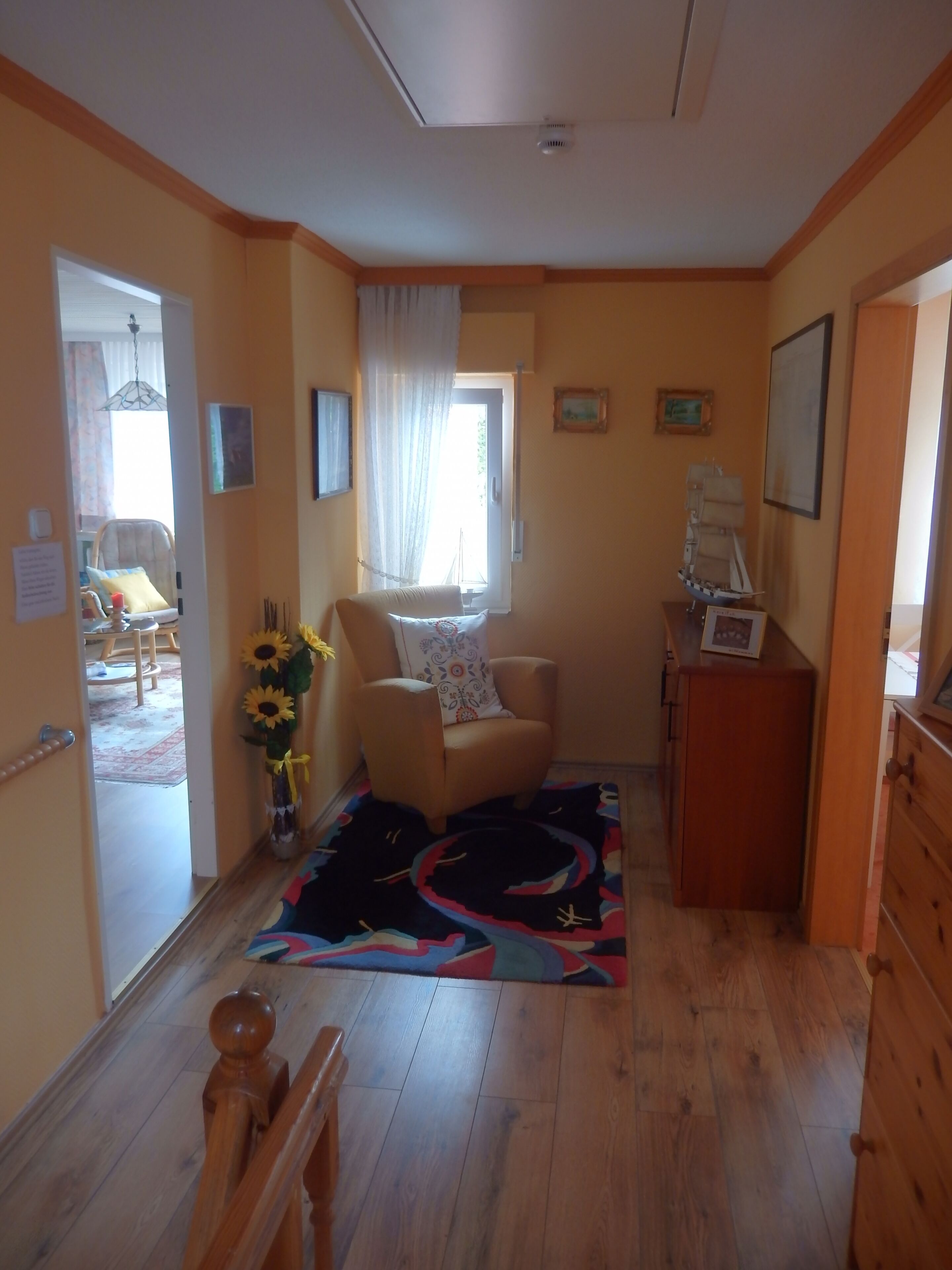 Ein gemütliches Wohnzimmer mit Holzboden, einem gemusterten Teppich, einem bequemen Sessel, einem kleinen Tisch mit einer Vase voller Sonnenblumen und einem Fenster mit Vorhängen.