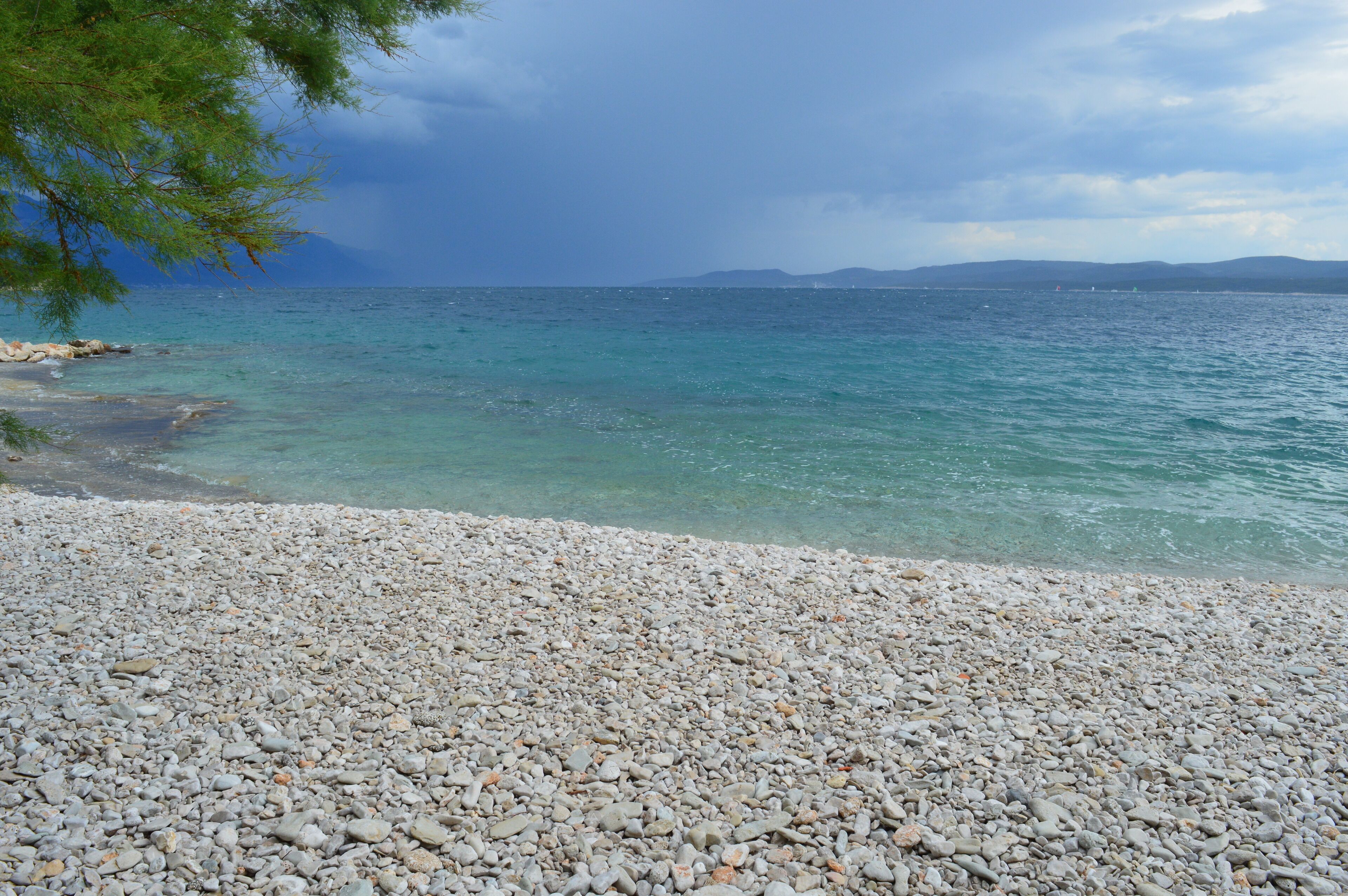 A pebbled beach with clear turquoise water and distant mountains under a cloudy sky.
