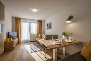 Uma sala de estar moderna com um sofá, uma mesa de jantar posta para dois, uma TV sobre um móvel de madeira e uma janela grande com cortinas.