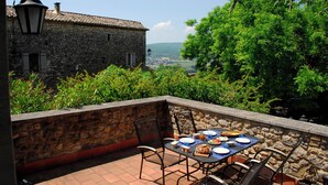 An outdoor patio with stone walls, a table set for dining, and a view of a stone building and greenery. - Bright duplex apartment with terrace and beautiful views (Cornillon)