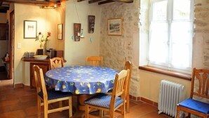 A dining area with a round table, wooden chairs, and a blue tablecloth. There is a window with a view of the outside, a radiator, and stone walls. - Bright duplex apartment with terrace and beautiful views (Cornillon)