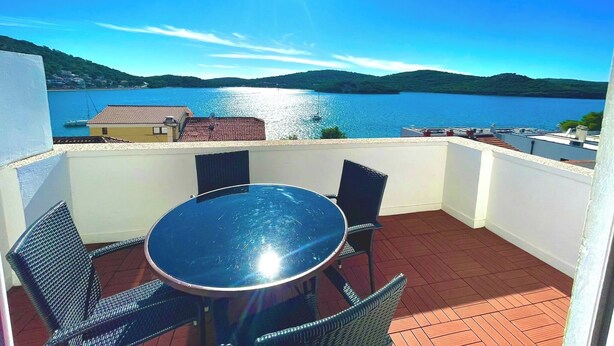 Eine Terrasse mit rundem Tisch und Stühlen, mit Blick auf ein Gewässer und Hügel.