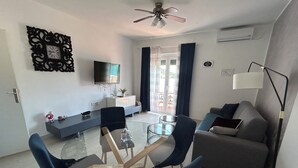 Ein modernes Wohnzimmer mit einem Glastisch, blauen Stühlen, einem grauen Sofa und einem Flachbildfernseher an der Wand.