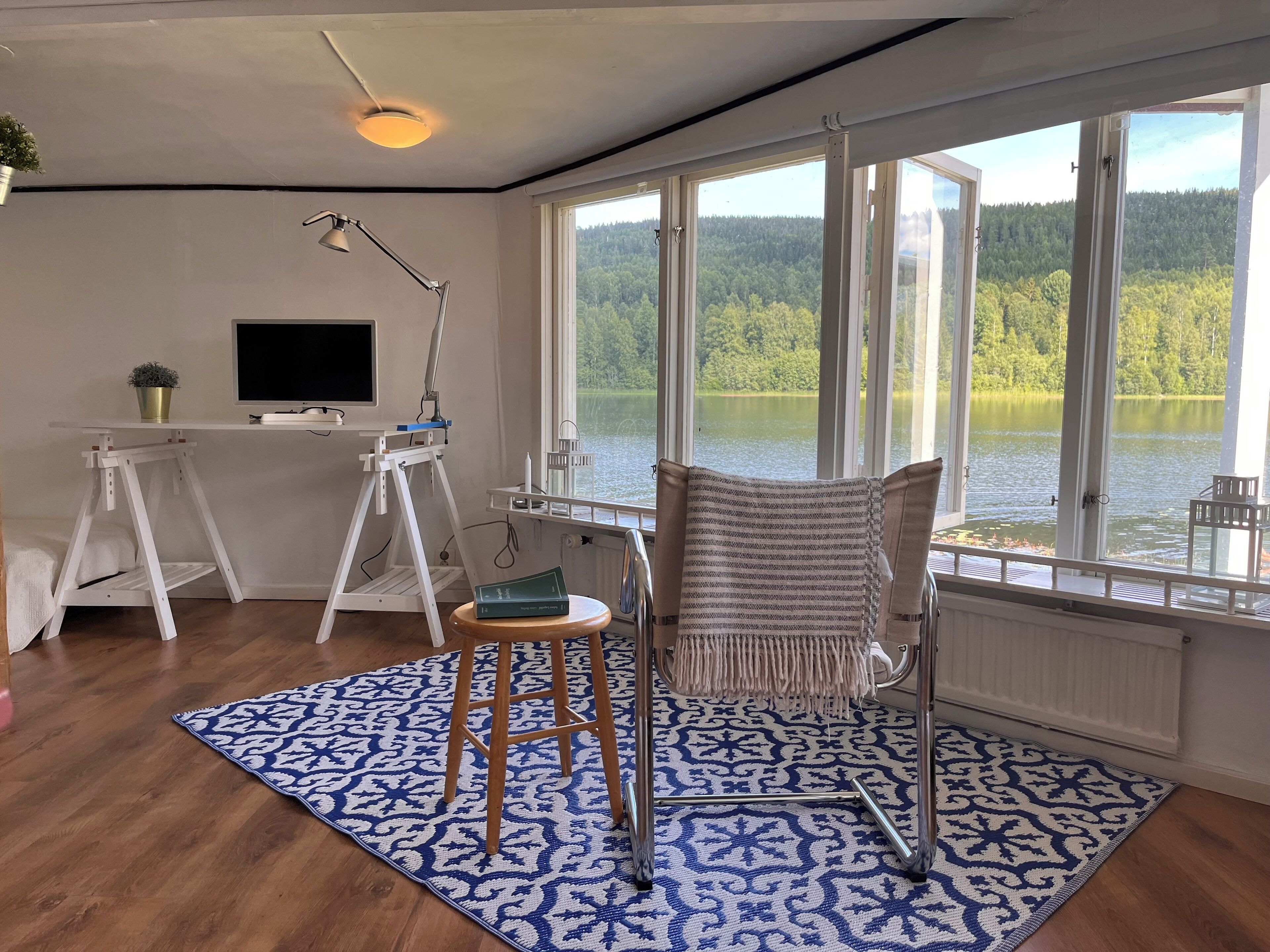Una habitación con un escritorio de madera, una silla, un ventanal con vistas a un lago y un bosque, y una alfombra estampada.