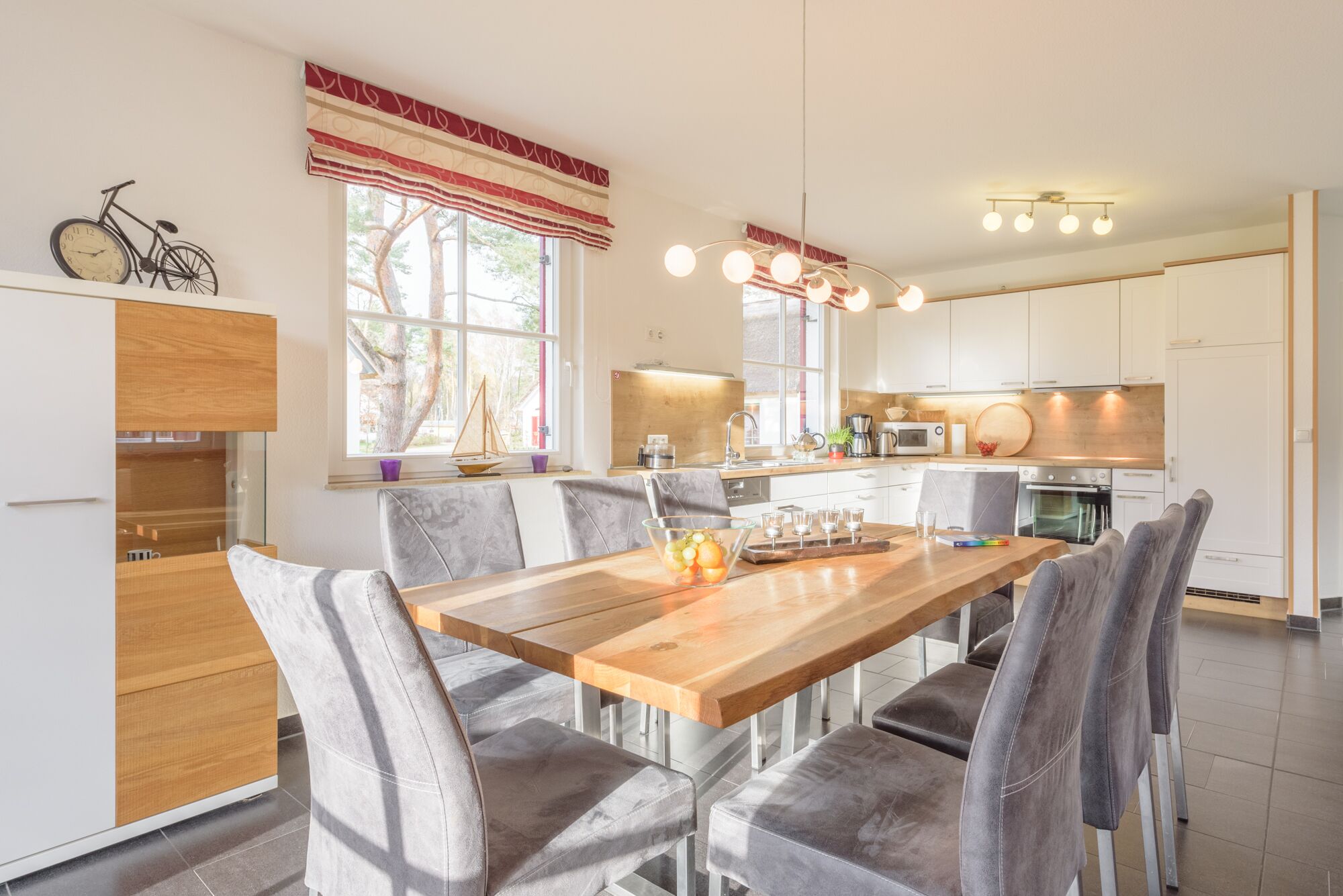 A modern kitchen with a wooden dining table, grey upholstered chairs, and a large window with a view of trees.