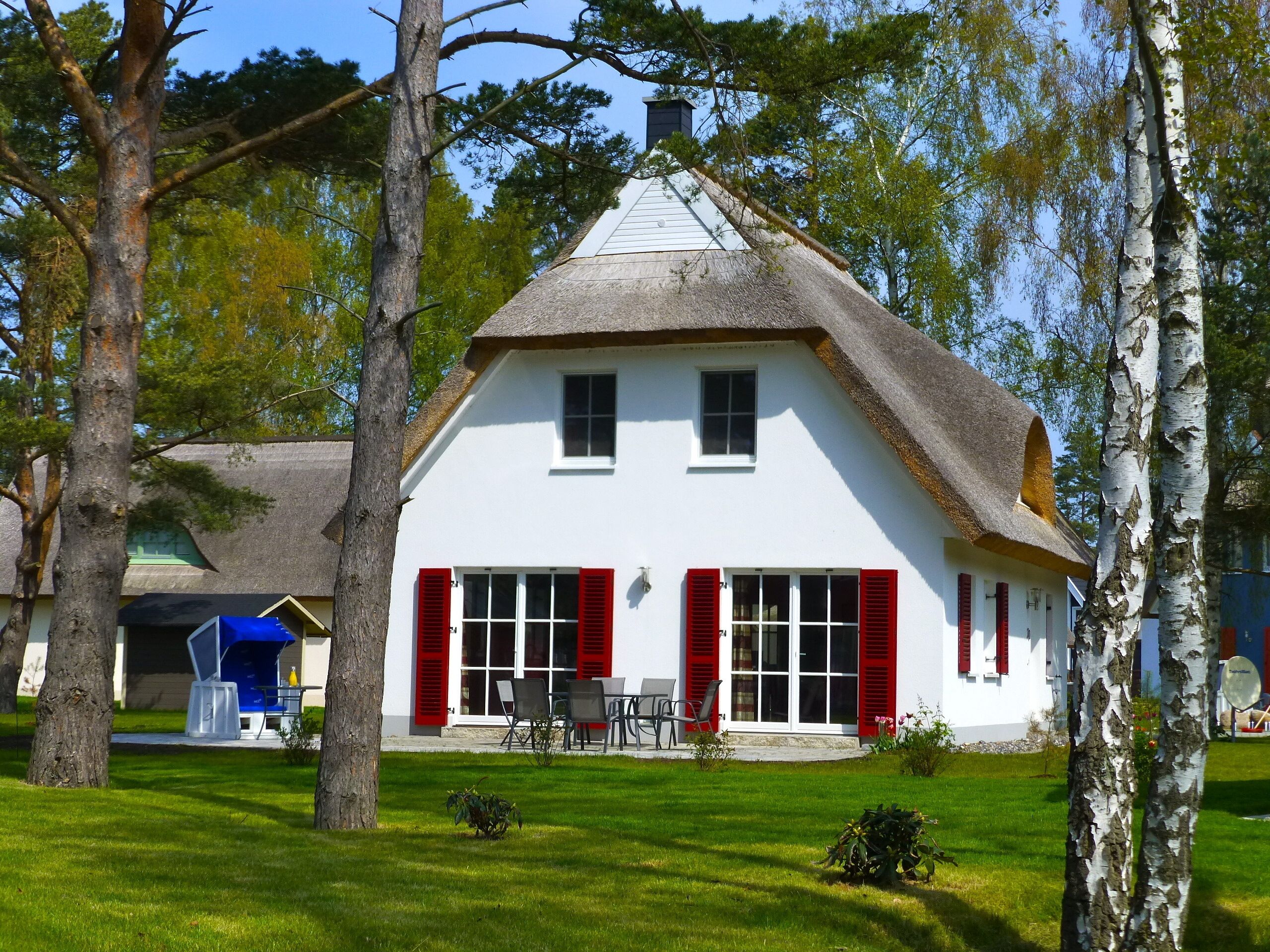 Ein weißes Haus mit roten Fensterläden, einem Strohdach und einem kleinen Garten mit Rasen und Bäumen.