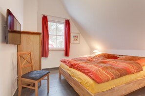 Ein Schlafzimmer mit Bett, Sessel, Fenster mit roten Vorhängen, Wand-Fernseher und einem gerahmten Bild.
