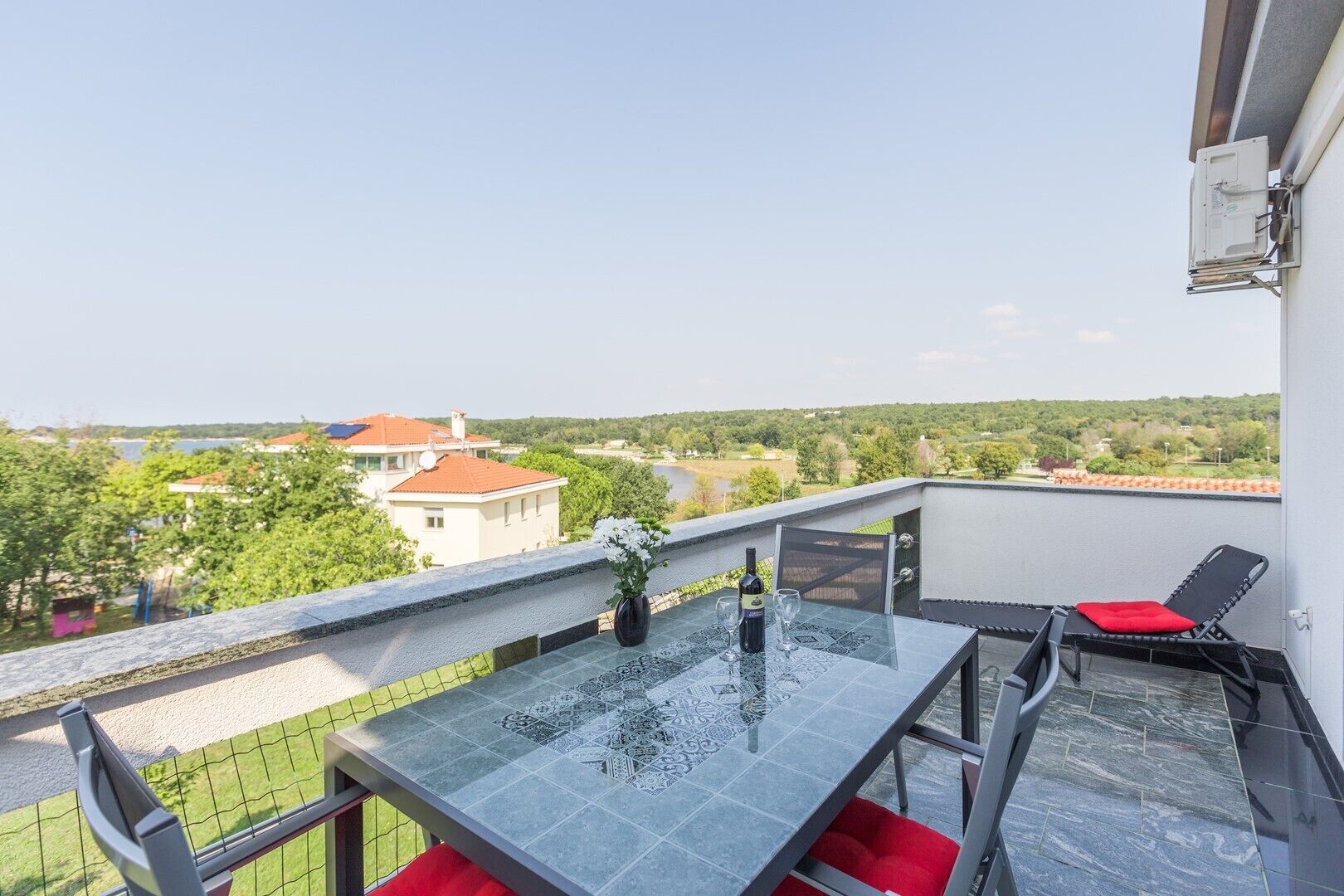 A balcony with a glass table, red cushions, and a bottle of wine.