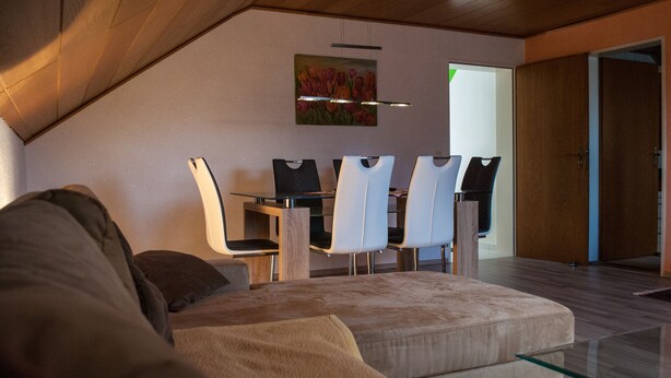 Een moderne woonkamer met een glazen salontafel, een beige bank en een eethoek met een houten tafel en witte stoelen.
