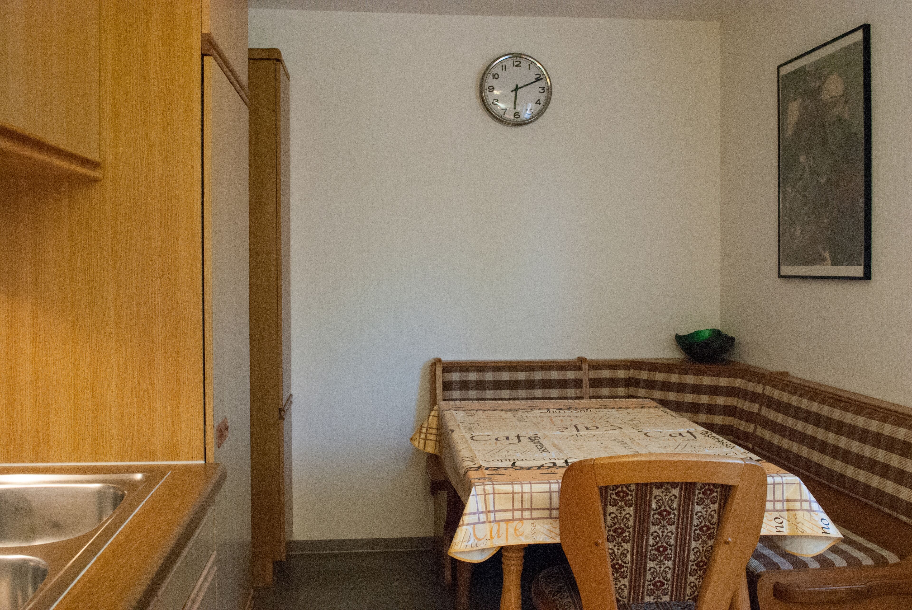 A kitchen with a wooden cabinet, a sink, a dining table with a checkered tablecloth, and a clock on the wall.