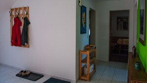 Een gang met een kapstok waarop een rode en een zwarte jas hangt, een houten plank met een mand, en een deur die naar een andere kamer leidt.