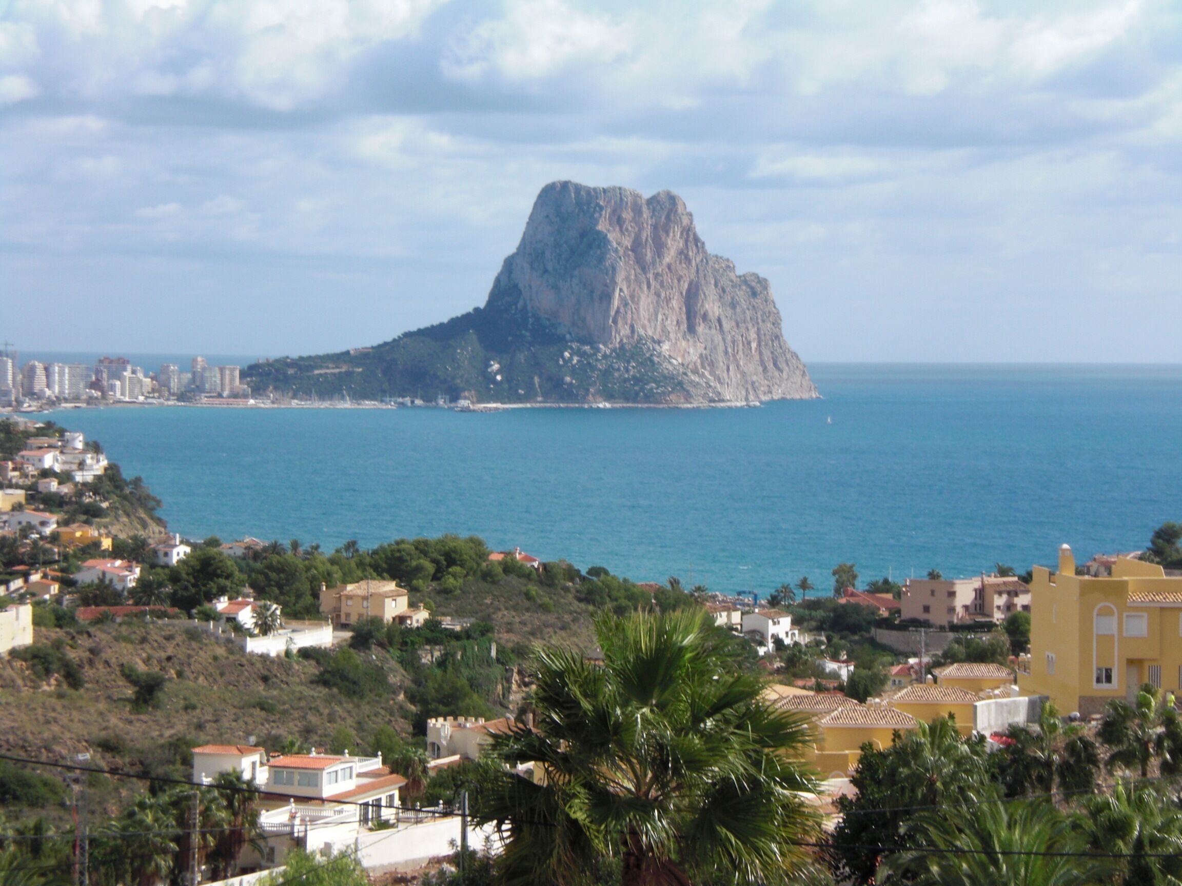 A coastal town with a prominent rocky hill in the background, a clear blue sea, and residential buildings with red-tiled roofs.