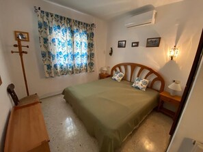 A bedroom with a bed, a nightstand, a lamp, a wall-mounted air conditioner, and a window with floral curtains. - Holiday house for 4 people with private pool and panoramic views (Calp)