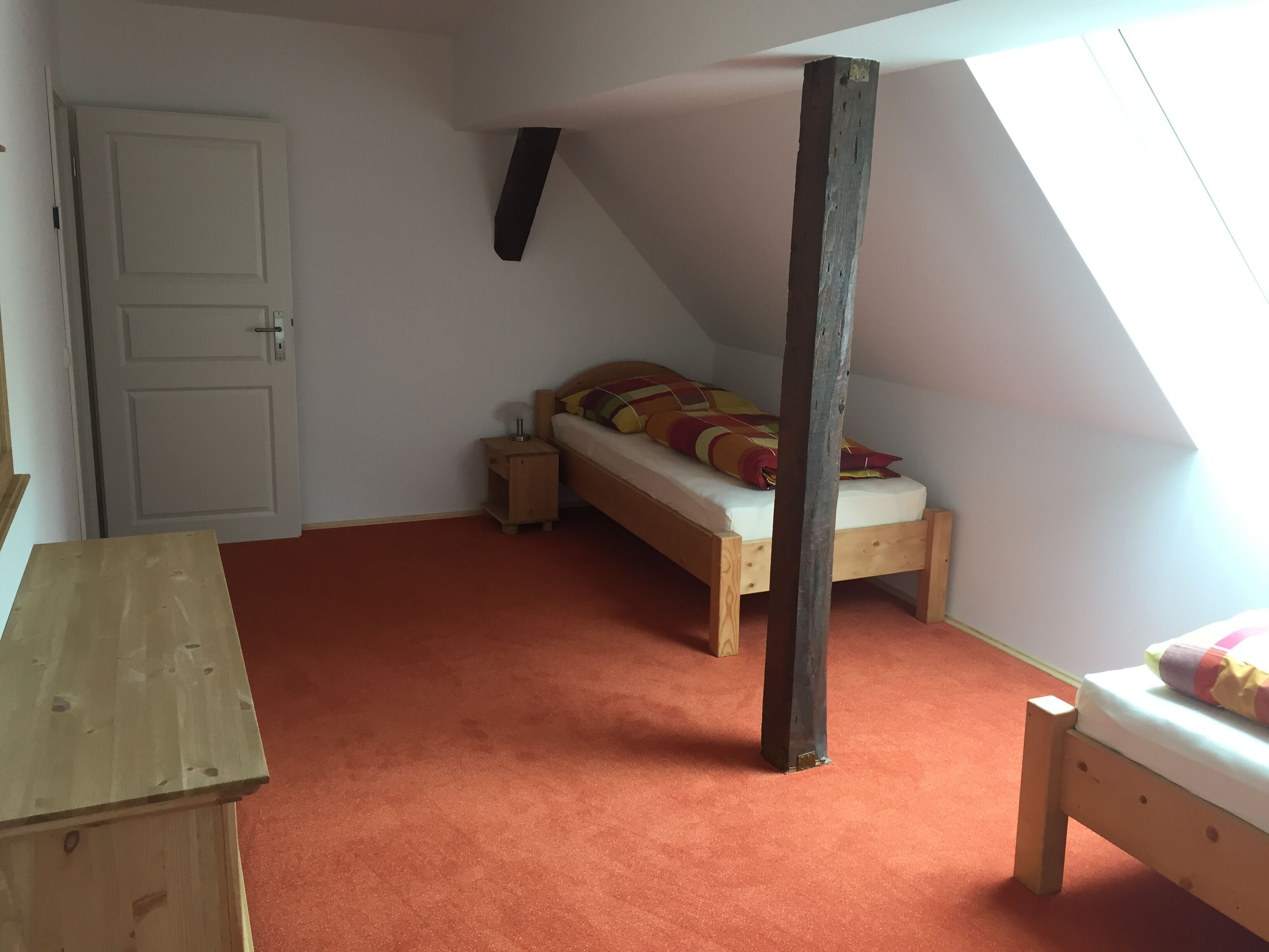 Une petite pièce accueillante avec un plafond en pente, un lit en bois avec une literie colorée, une petite table de chevet en bois et une porte.
