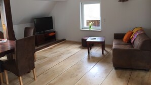 Un salon avec un plancher en bois, un téléviseur posé sur un meuble, une fenêtre avec des rideaux, un canapé avec des coussins et une petite table.