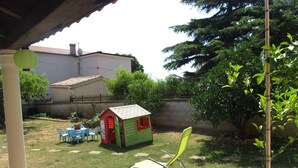 Un giardino sul retro con una casetta da gioco, una sedia verde e un vaso con fiori rossi.