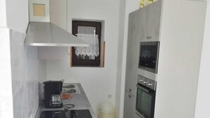 Cucina compatta con fornello, forno e microonde. Si nota una piccola finestra con tenda, e un cestino per i rifiuti.