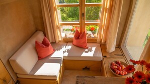 A cozy room with a wooden bench and cushions, a window with curtains, and a basket of fruit on a small table.