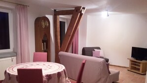 Ein Essbereich mit einem runden Tisch und Stühlen, ein Holzstuhl mit Rückenlehne, ein Fernseher auf einem Holzboden und ein Fenster mit Vorhängen.