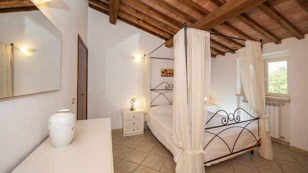 Un dormitorio con cama con dosel, mesita de noche, florero y una ventana con cortinas. El techo presenta vigas a la vista.
