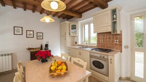Una cocina con armarios blancos, estufa a gas, lavabo y ventana. Hay una mesa con cesta de frutas y flores.