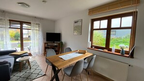 A modern living room with a dining area, large windows, and a flat-screen TV. - Hunsruecklust vacation apartment with e-bike rental (Beltheim)