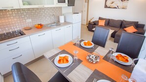 Uma cozinha moderna com armários brancos, micro-ondas e pia. Há uma área de jantar com uma mesa posta para dois, jogos americanos laranja e uma tigela de frutas.