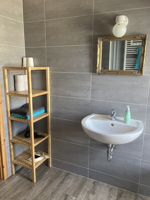 Un bagno con uno scaffale in legno contenente asciugamani e articoli da toilette, un lavandino bianco con un rubinetto e uno specchio con cornice decorativa.