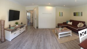 A living room with a sofa, coffee table, TV, and a door leading to another room. - (g) Roosike vacation in Bacharach-Steeg, modern apartment in the countryside (Bacharach)
