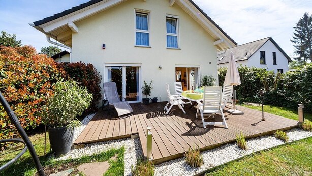 Ein zweistöckiges Haus mit einer Holzterrasse, einer Terrasse mit Gartenmöbeln und einem Garten mit verschiedenen Pflanzen.