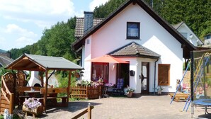 A two-story house with a wooden porch, a chimney, and a small garden area with flowers and a wooden bench. - Pure relaxation with steam shower and whirlpool in a comfort cottage  (Bad Berleburg)