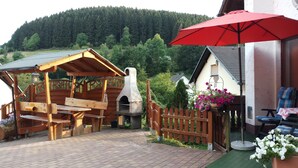 A wooden outdoor seating area with a brick oven, a wooden fence, and a red umbrella. - Pure relaxation with steam shower and whirlpool in a comfort cottage  (Bad Berleburg)